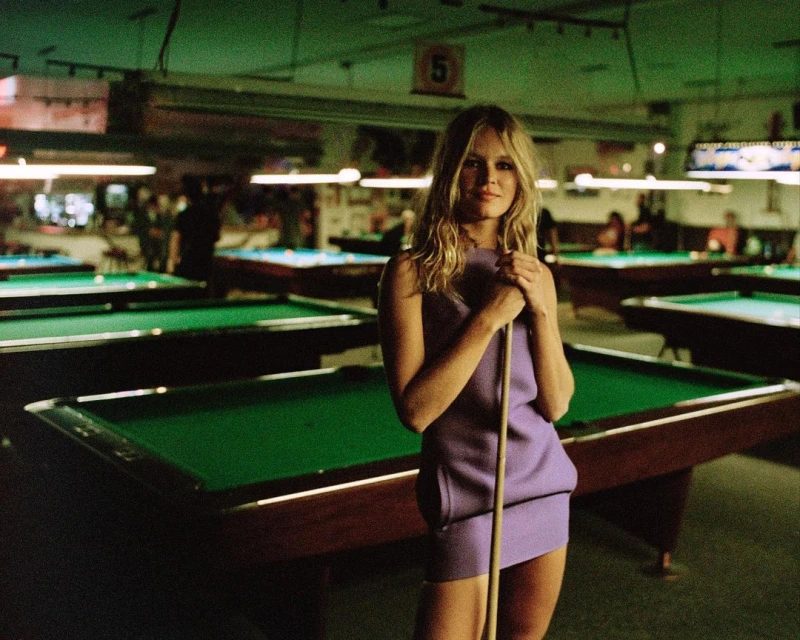 Woman in purple dress poses with pool cue stick in a billiards room with green pool tables and soft lighting, capturing a casual, relaxed atmosphere.