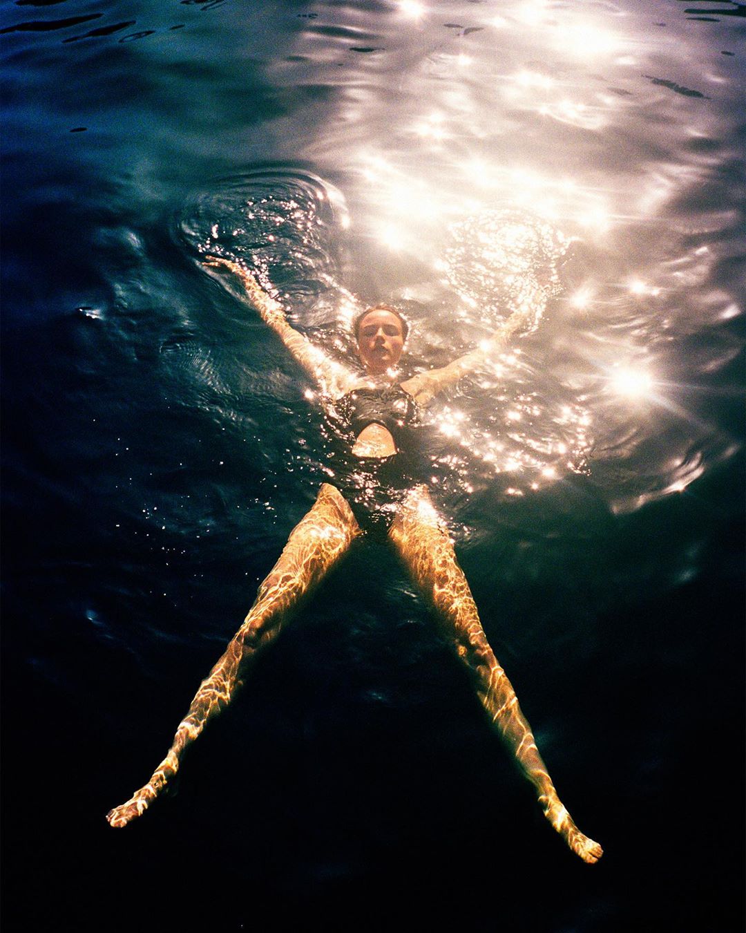 Woman in a black swimsuit floats on her back in dark water with her arms and legs outstretched as the sun shines brightly overhead.