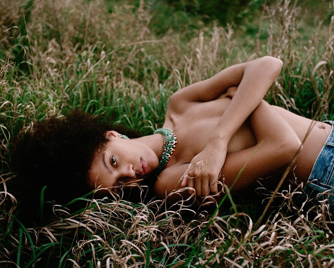 A young woman with dark curly hair lies on her side in a field of tall grass, wearing a blue jeans and a turquoise statement necklace.