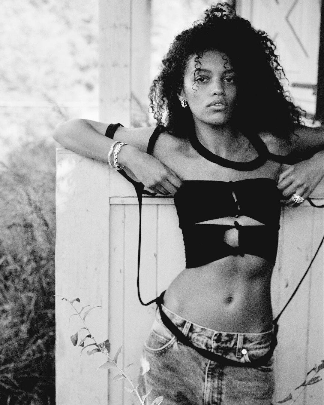 Black and white portrait of a woman with curly hair wearing a modern black top and jeans posing outdoors, leaning against a white surface.