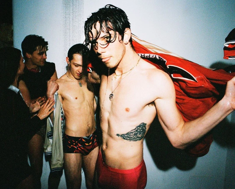 Wet, shirtless male athlete in swimwear stands with teammates indoors. Athletic gear and wet skin add to the raw, sporty atmosphere.