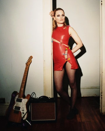 Blonde woman in a red dress poses near an electric guitar and amp in a doorway against a white wall, creating a striking portrait.
