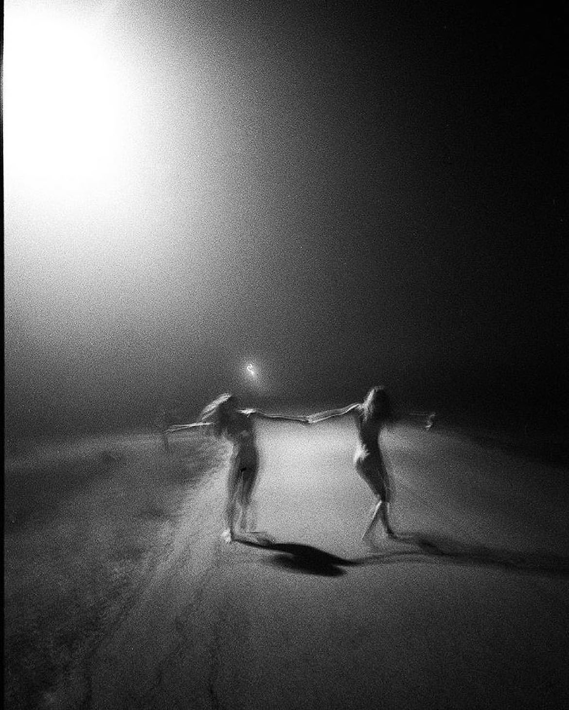 Two women holding hands, seemingly dancing, in a grainy black and white photograph with a bright light source creating strong contrast.