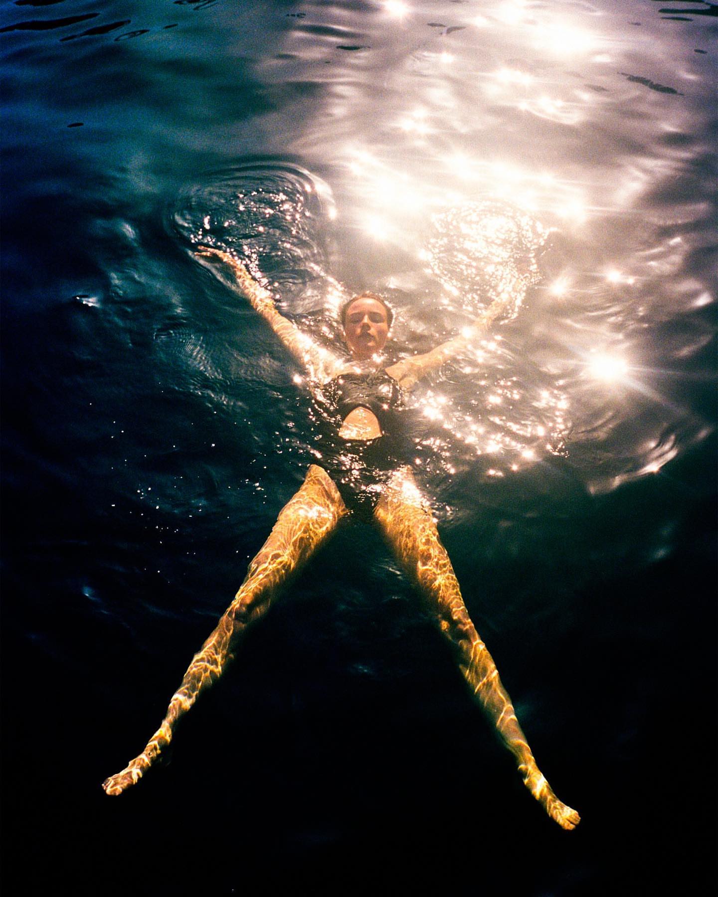 Woman in a black swimsuit floats in dark water with arms outstretched, illuminated by sunlight creating a high-contrast and serene scene.