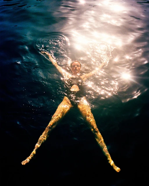 Woman in a black swimsuit floats in dark water with arms outstretched, illuminated by sunlight creating a high-contrast and serene scene.
