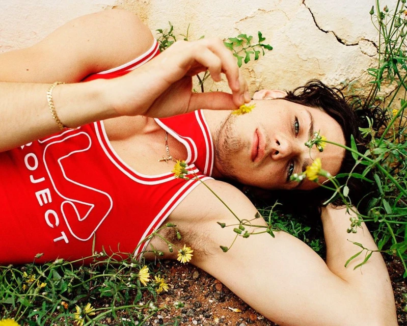 An adult male model lies on a bed of greenery and tiny yellow flowers, wearing a red and white tank top with a gold bracelet and necklace.