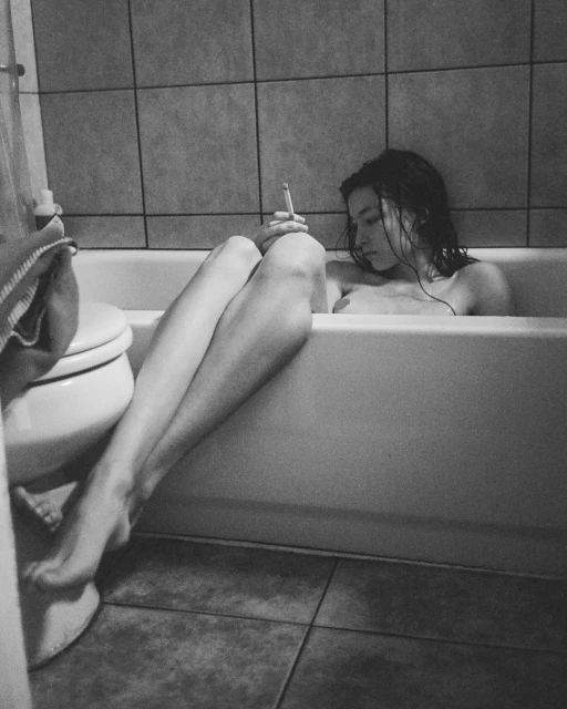 A woman relaxes in a tiled bathroom tub, holding a cigarette. She has dark hair, eyes closed, legs extended toward the toilet.