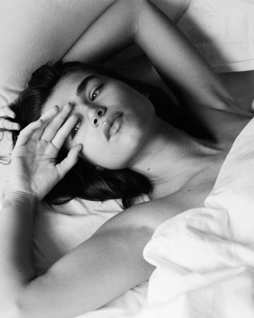 Black and white portrait of a woman partially covering her face with her hand while lying in bed with white sheets, looking at the camera.