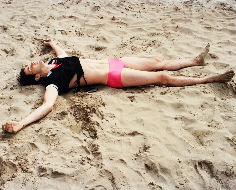 Athletic man lying on sandy beach in pink swimsuit and black crop top with arms outstretched, enjoying the sun and ocean breeze.