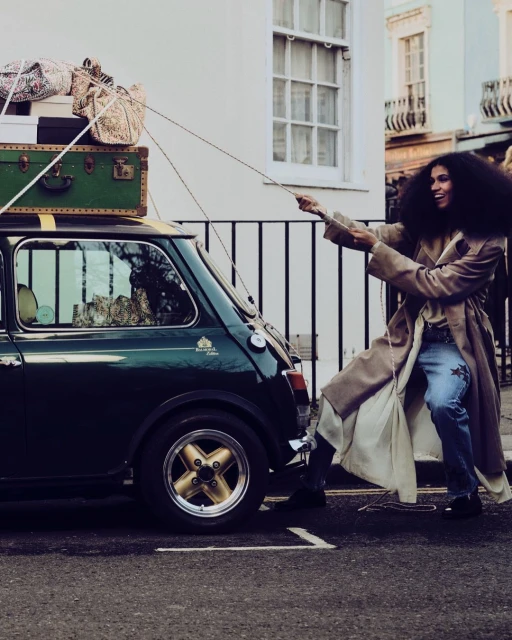 A stylish adult female pulls a vintage green Mini Cooper with luggage on top, parked on a city street near white buildings and a black fence.