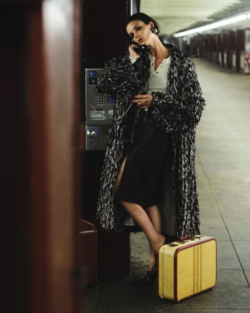 Fashion shot of a woman in a patterned coat using a vintage payphone in a subway station with a yellow suitcase at her feet.