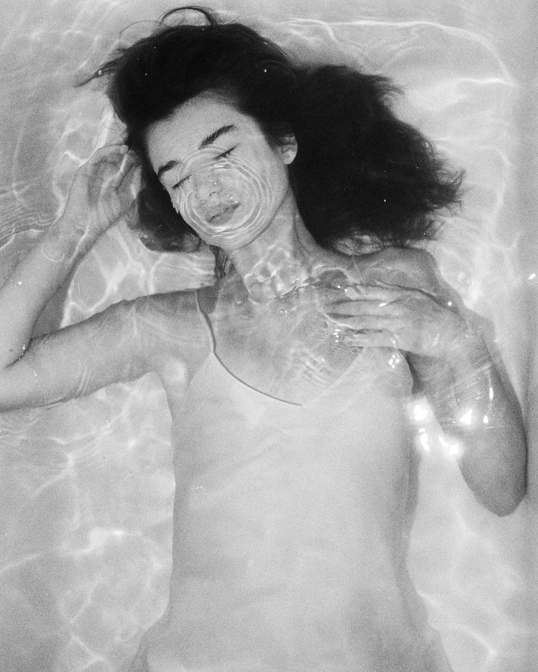 Monochrome portrait of a woman with closed eyes, lying in water, creating a serene and reflective atmosphere with ripples on the water's surface.