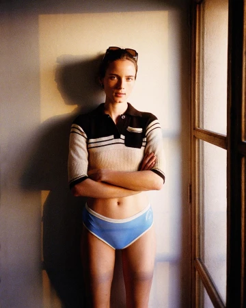 Woman with arms crossed wearing a striped polo top and blue briefs standing indoors near a window by photographer Bryan Liston.