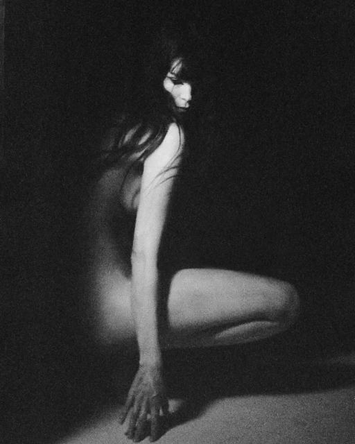 Monochrome portrait of a woman with long dark hair in a crouched position, set against a dark background, with soft, diffused lighting.