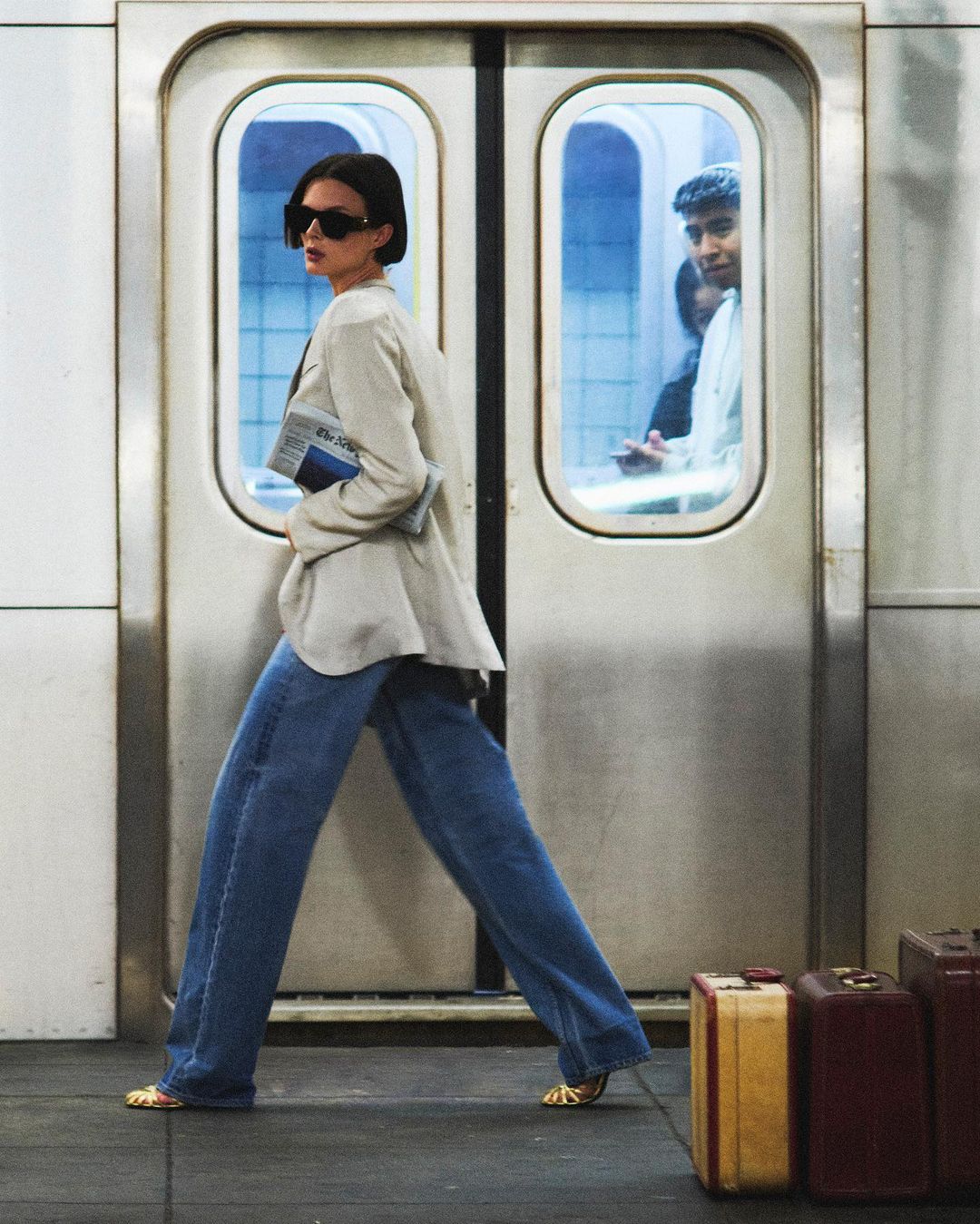 Fashionable woman in oversized blazer and wide-leg jeans walks past subway doors and vintage luggage on a platform with soft light.