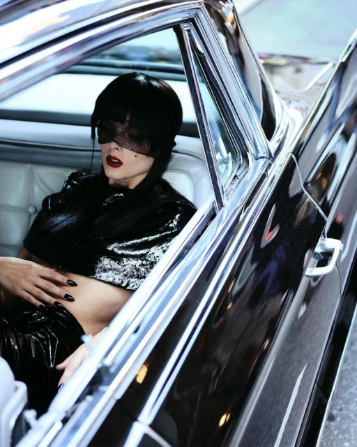 Woman in a car wearing black clothing and a net veil. The car has a chrome exterior and white leather interior.