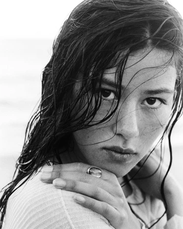 Close-up portrait of a woman with wet dark hair framing her face, resting her hand with a ring near her chin.