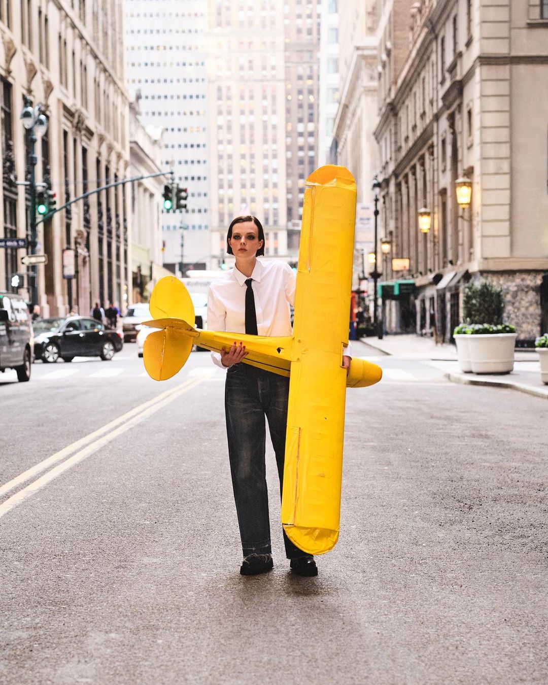 Woman in a white shirt and dark jeans stands on a city street holding a large yellow airplane toy, buildings and traffic in background.