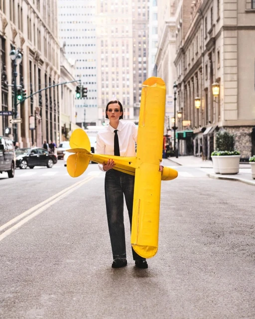 Woman in a white shirt and dark jeans stands on a city street holding a large yellow airplane toy, buildings and traffic in background.