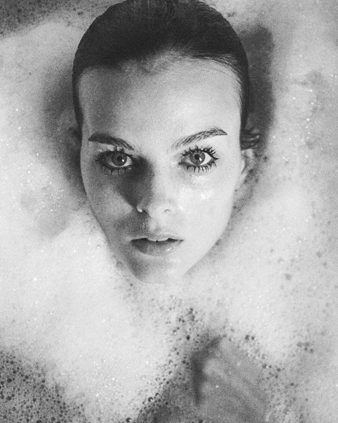 Black and white portrait of a young woman immersed in a bubble bath, her face and shoulders visible amidst the foamy water.