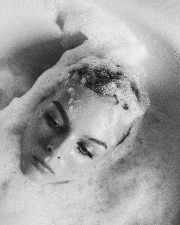 Black and white portrait of a woman submerged in a bathtub surrounded by soapy foam, photographed by Bryan Liston