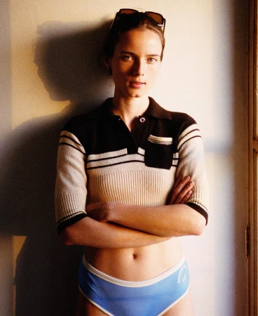 Woman in short-sleeve polo sweater and blue briefs leaning against a wall with sunglasses on her head by Bryan Liston