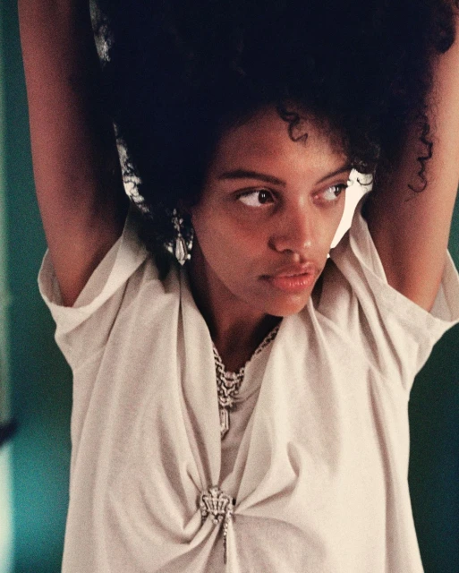 Close-up of a stylish African-American woman with dark hair posing indoors wearing a white dress, silver necklace, and earrings.