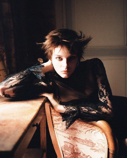A young adult woman with short, tousled hair in a lace top poses in a dimly lit interior room next to a patterned antique chair.