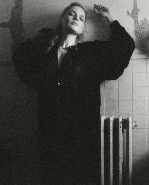Monochrome portrait of a blonde adult woman in a dark top leaning against a tiled wall near a radiator in an indoor setting.