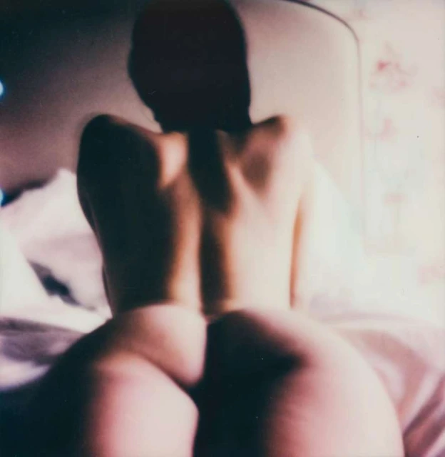Back view of a nude woman sitting on a bed in a photograph with a blurry effect, soft lighting and pink and white colors.