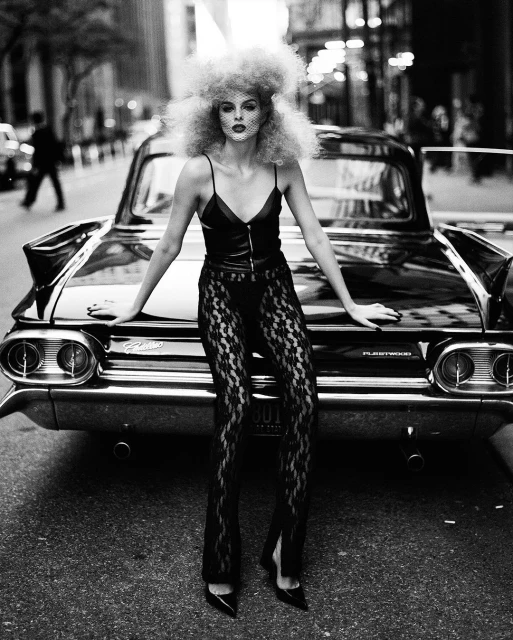 Black and white fashion shot of a woman in lace pants leaning on a vintage car in the city, showcasing edgy style and urban elegance.
