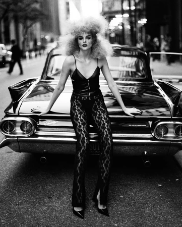 Black and white fashion shot of a woman in lace pants leaning on a vintage car in the city, showcasing edgy style and urban elegance.