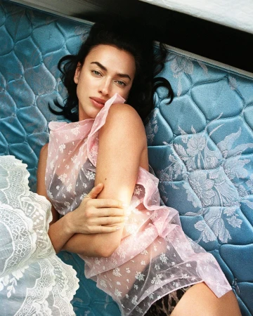 Woman in a pink ruffled top poses on a blue floral mattress with a white lace coverlet in a fashion photography editorial. 