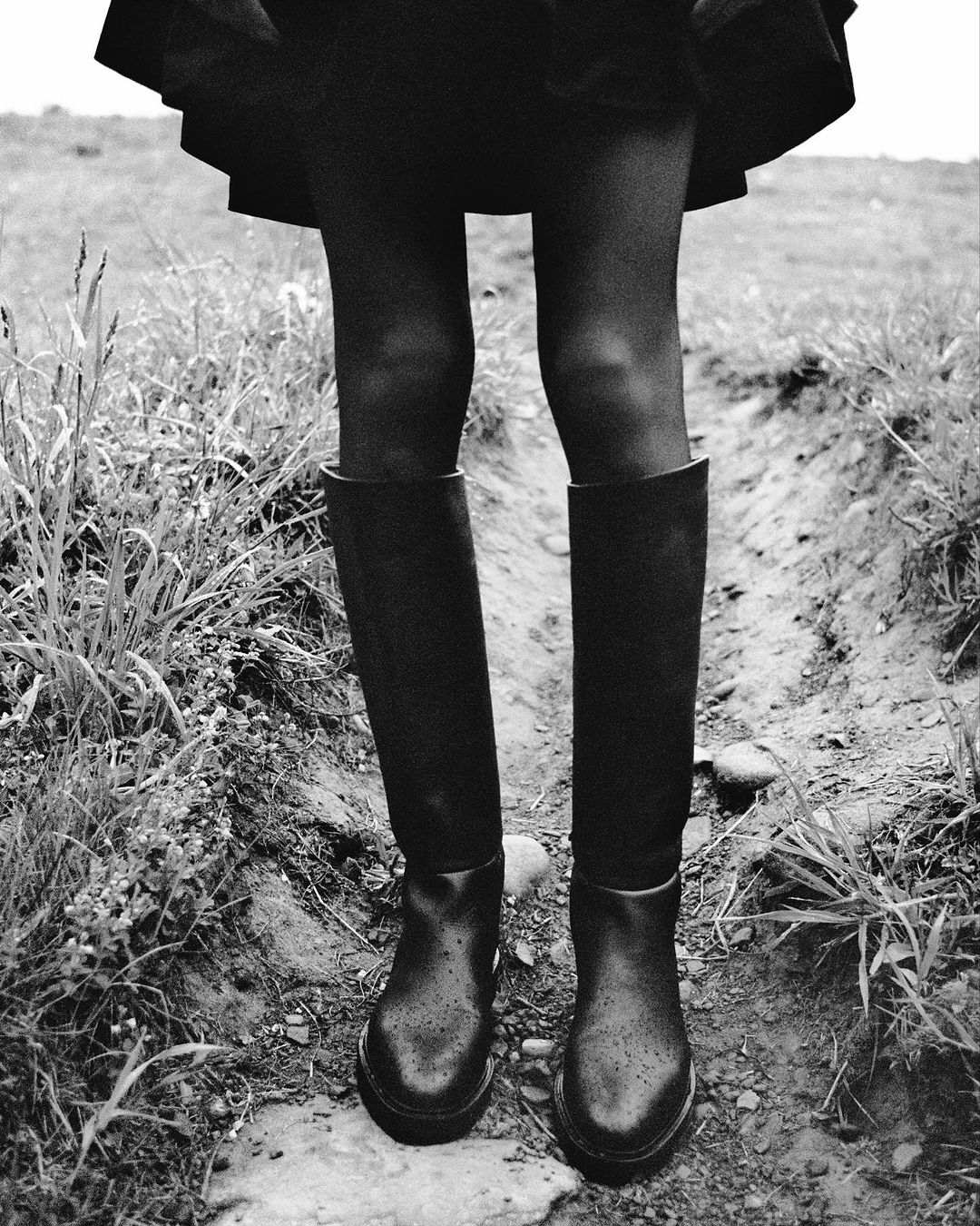 Monochrome shot of a person wearing black tights and boots, standing on a dirt path surrounded by tall grass in a field.