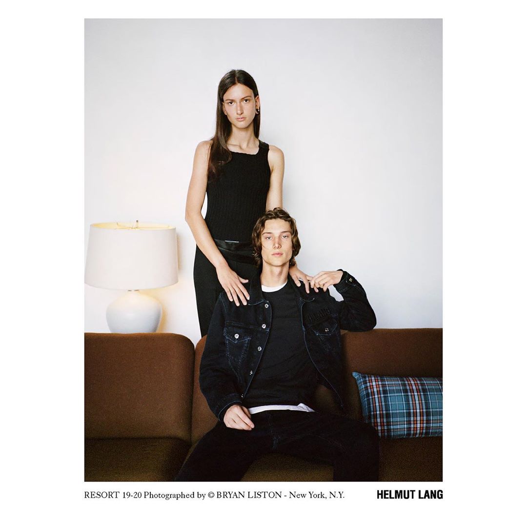 A stylish studio portrait of a young adult couple. The woman stands behind the seated man, both dressed in fashionable black clothing.