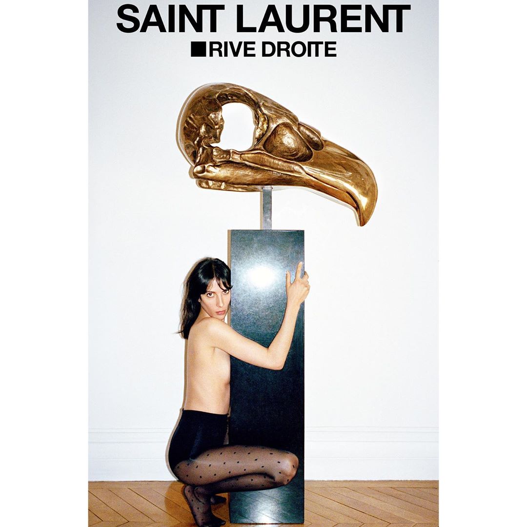 Gold bird skull sculpture, displayed with a crouching brunette adult woman in black tights in a studio setting for Saint Laurent Rive Droite.