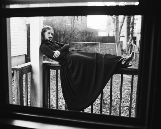 Monochrome portrait of a woman in a dark dress leaning against a wooden porch railing, shot through a window in New York.