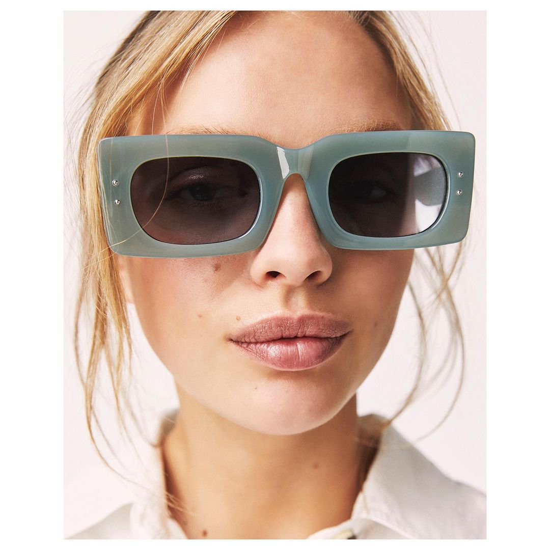 Close-up of a blonde adult woman wearing chunky, sage green sunglasses. The image is brightly lit with a white background.
