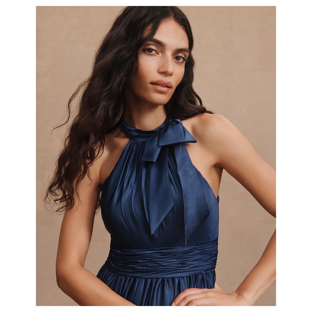 Woman wearing a navy blue halter dress with a bow stands indoors against a neutral backdrop in a fashion-style portrait.