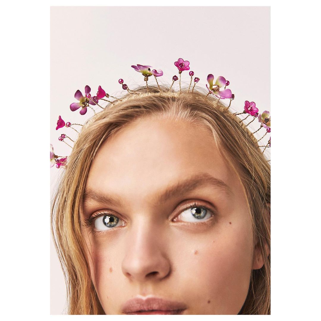 Close-up of a young woman with light hair and blue eyes, wearing a flower crown with purple and pink blossoms on a neutral background.