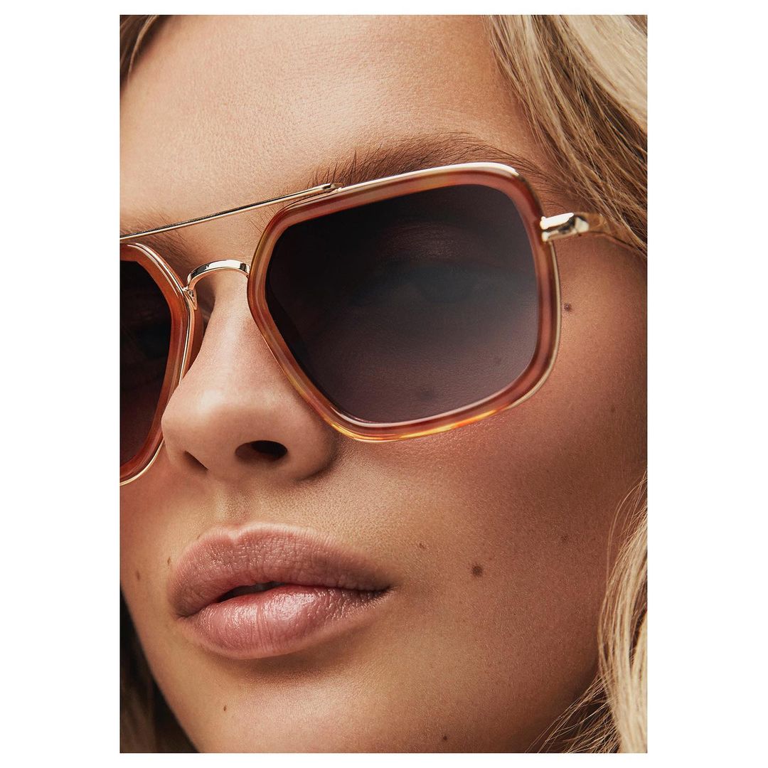 Close-up of a blonde adult woman wearing square aviator sunglasses with gold frames and a dark gradient lens; beauty, fashion, lifestyle.