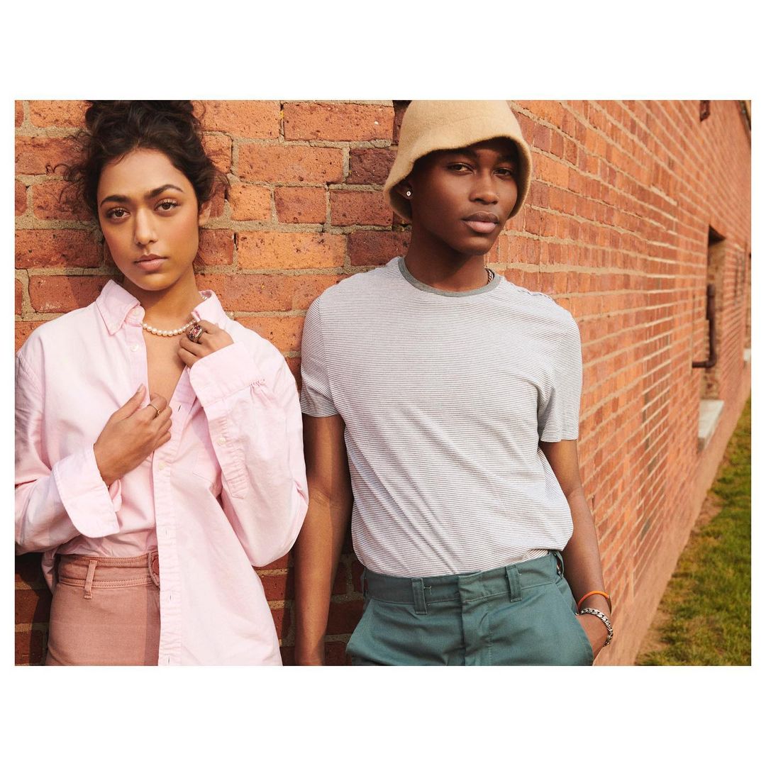 Two fashion models pose against a brick wall wearing casual clothing in a medium shot with shallow depth of field and natural lighting.
