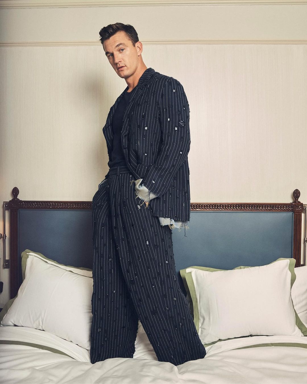 A stylish adult male stands on a bed in a fashionable striped suit and dark shirt, posing indoors in a portrait framed by a headboard and pillows.