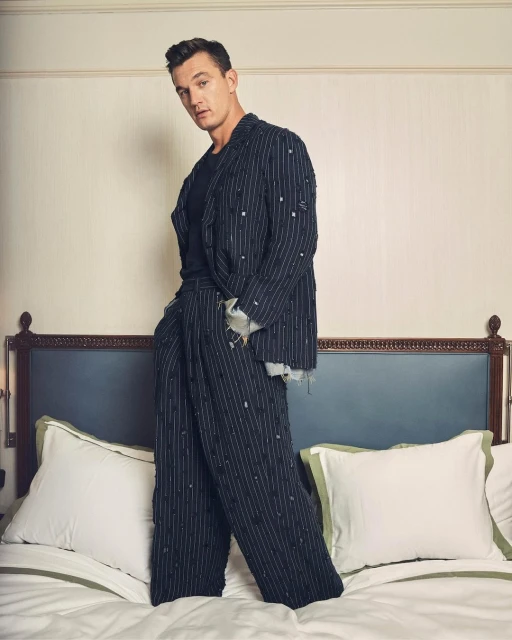 A stylish adult male stands on a bed in a fashionable striped suit and dark shirt, posing indoors in a portrait framed by a headboard and pillows.