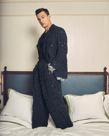 A stylish adult male stands on a bed in a fashionable striped suit and dark shirt, posing indoors in a portrait framed by a headboard and pillows.