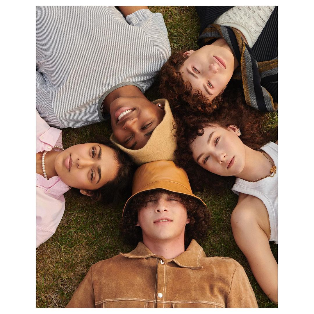 A diverse group of five young adults lies on the grass in a starburst pattern, wearing fashionable casual clothing and hats and looking at the camera.