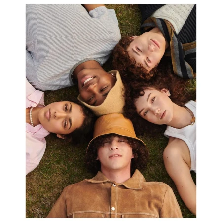 A diverse group of five young adults lies on the grass in a starburst pattern, wearing fashionable casual clothing and hats and looking at the camera.
