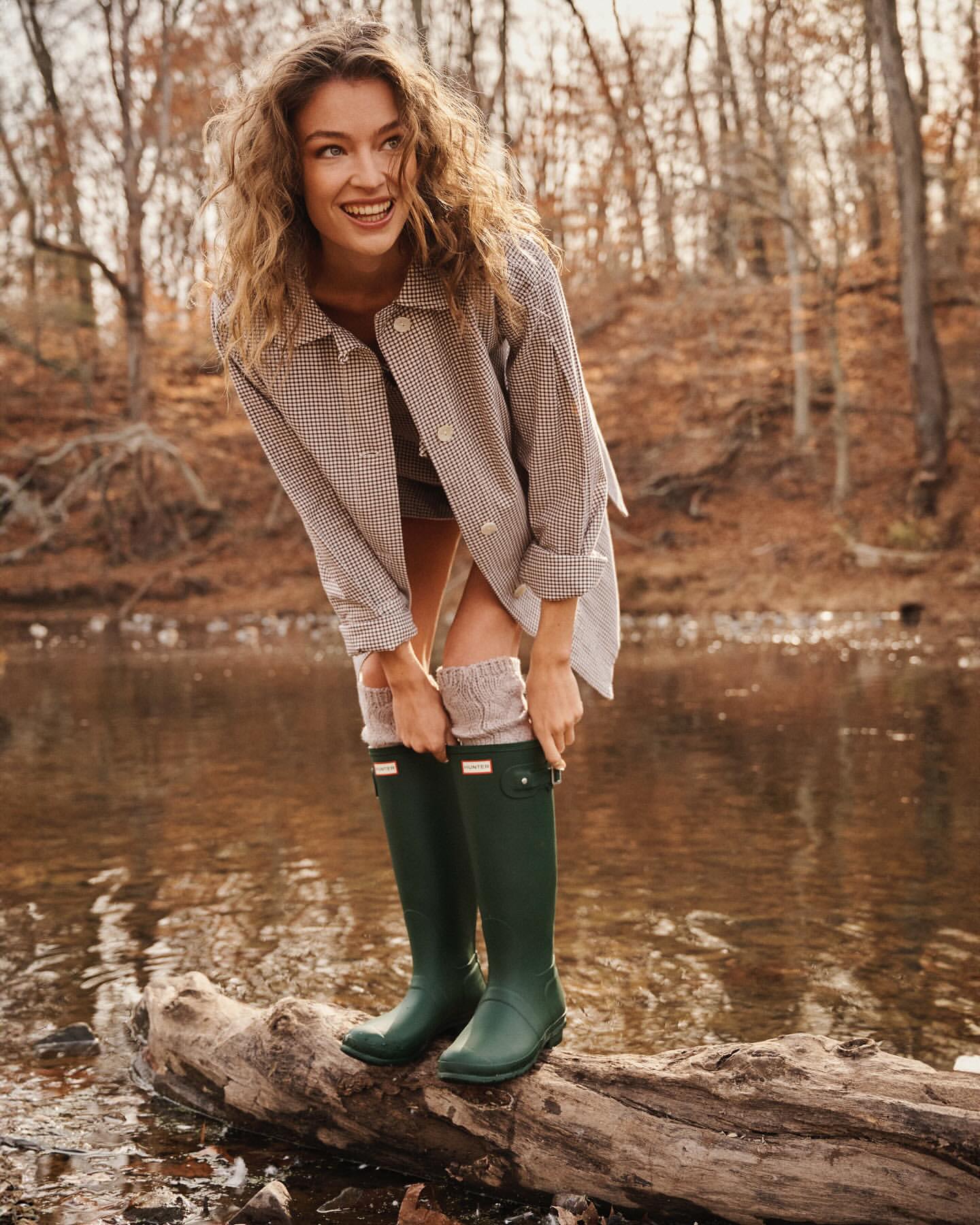 Smiling woman in green rubber boots poses on a log by a river surrounded by autumn trees in an outdoor setting.