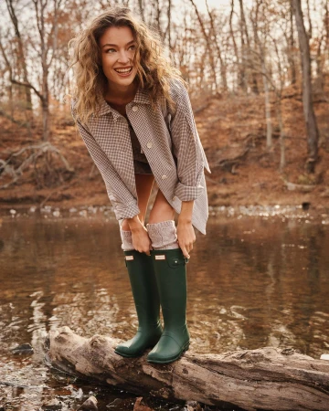 Smiling woman in green rubber boots poses on a log by a river surrounded by autumn trees in an outdoor setting.
