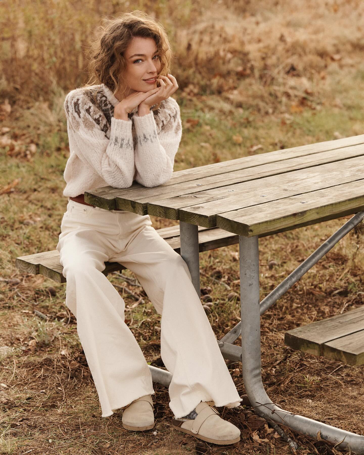 A woman sits at a wooden picnic table outside, wearing a fair isle sweater, cream-colored jeans, and clogs. She rests her chin on her hands.